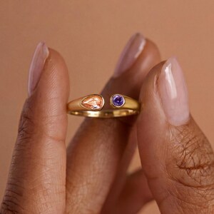 May include: A gold open-style ring featuring two distinct gemstones. One end of the band holds a pear-shaped peach-colored stone, while the other end features a small, round, vibrant purple stone. The metal band has a smooth, polished finish.