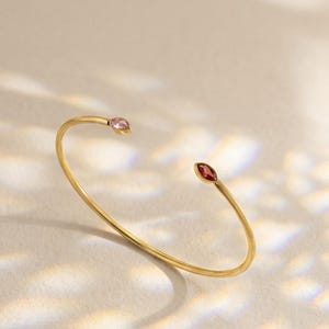 May include: A gold-toned open cuff bracelet featuring two marquise-cut stones at the ends. One stone is a light pink colour, while the other is a deep red. The bracelet has a thin, smooth metal band and is positioned to show its open design.
