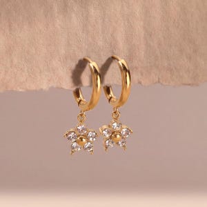 May include: A pair of gold hoop earrings featuring dangling star-shaped charms. Each charm is adorned with five clear, sparkling stones arranged in a star pattern with a small gold centre. The earrings have a polished finish and a hinged closure.