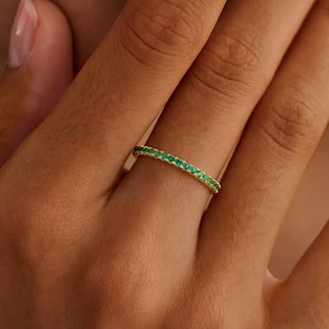 Puede incluir: Un anillo de oro con una banda de pequeñas y vibrantes piedras verdes. El anillo se muestra en una mano, mostrando el delicado diseño y el brillo de las piedras similares a esmeraldas. El anillo es una joya.