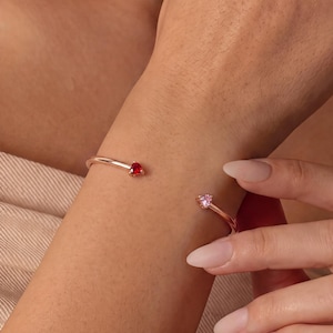 May include: A rose gold open cuff bracelet featuring two small, round-cut gemstones at the ends. One gemstone is a deep red colour, while the other is a light pink colour. The metal band is thin and smooth, encircling the wrist with an open gap.