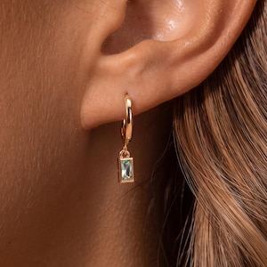 May include: A gold hoop earring featuring a small, rectangular, light blue baguette-cut gemstone charm dangling from the bottom. The earring is secured through the earlobe, showcasing a polished metallic finish and a clear, pale blue stone.