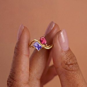 Könnte beinhalten: Ein goldener Ring mit einem überlappenden Banddesign und zwei markanten Edelsteinen. Ein Stein ist ein rechteckig geschliffener hellvioletter Edelstein, der andere ein birnenförmiger, leuchtend pinker Stein. Das Metallband hat eine glatte Oberfläche.