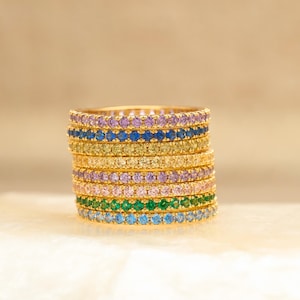May include: A stack of gold-toned rings, each adorned with a row of colorful gemstones. The rings display a spectrum of colors, including blue, green, yellow, purple, and pink. The rings are stacked on a white surface against a soft, neutral background.
