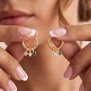 May include: A pair of gold hoop earrings, each featuring three small dangling charms with round-cut stones in pink, clear, and green. The earrings are polished gold metal, and the stones are set in small prongs, creating a delicate and symmetrical look.