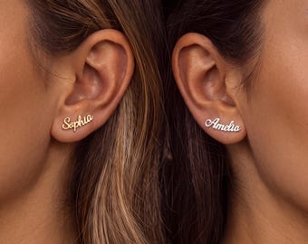 May include: Two stud earrings featuring custom name designs in a cursive script font. One earring is gold-toned and displays the name Sophia, while the other is silver-toned and displays the name Amelia, both worn on the earlobe.