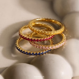 Peut inclure: Un empilement de quatre bagues dorées, chacune dotée d'une bande continue de petites pierres précieuses rondes de couleur jaune, rouge, rose clair et bleue. Les bagues sont disposées en groupe, mettant en valeur la finition métallique et les pierres.