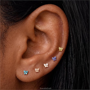 May include: An ear with five small butterfly stud earrings in a variety of colours, including blue, yellow, and clear, set in gold. The earrings are arranged in a line along the ear. The image is a close-up.