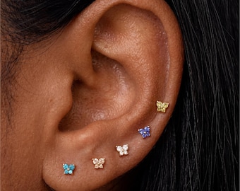 May include: Close-up of an ear adorned with five small butterfly stud earrings. The earrings are in various colors, including blue, yellow, and clear, and are set in gold. The earrings are arranged in a line along the ear.