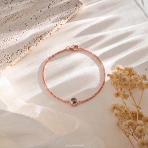 May include: Rose gold bracelet featuring a small, round, grey gemstone. The delicate chain is connected with a lobster clasp. The bracelet is displayed on a white, textured surface with dried flowers.