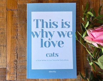 Pode incluir: Um livro azul claro com o título "This is why we love cats" em branco. O livro está sobre uma superfície de madeira, com uma rosa rosa e folhas verdes. O subtítulo diz "a love letter to our favorite furry divas."