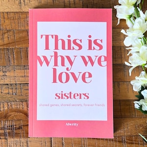 Op de afbeelding: Een roze boek met de tekst "This is why we love sisters" in een vetgedrukte, roze lettertype. Het boek ligt op een houten oppervlak, met witte bloemen op de achtergrond. De tekst hieronder luidt "shared genes, shared secrets, forever friends."