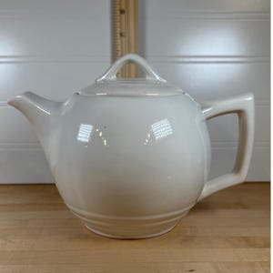 Puede incluir: Tetera de cerámica color crema con tapa y asa. La tetera tiene un cuerpo redondeado, un pico y un asa de bucle. La tapa tiene un asa pequeña. La tetera está sobre una superficie de madera.