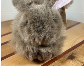May include: A plush rabbit toy covered in shaggy, light brown fur. The rabbit has two upright ears, one of which is lined with light pink fabric on the inner side. A small, tan-colored paper tag is attached to the side of the rabbit's head.