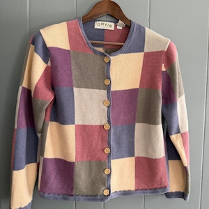 May include: A long-sleeve button-up cardigan featuring a multi-colored checkerboard pattern in shades of cream, dusty rose, purple, and slate blue. The garment has a round neckline, a front placket with six light brown buttons, and an Orvis brand label.