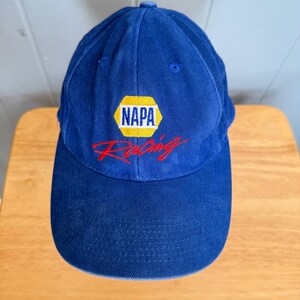 Pode incluir: Um boné de beisebol azul royal com um logótipo NAPA bordado dentro de um hexágono amarelo, com a palavra Racing cosida em letra cursiva vermelha por baixo. O boné tem uma copa estruturada, uma aba curva e costuras azuis a condizer.