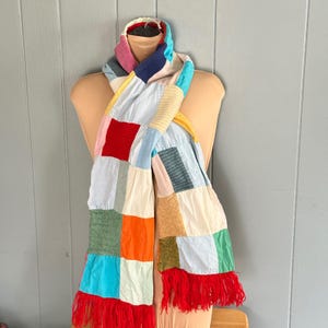 May include: A patchwork scarf with a variety of colorful fabric squares. The scarf has a red fringe at the ends and is displayed on a mannequin. The scarf's colors include red, blue, green, orange, and yellow.