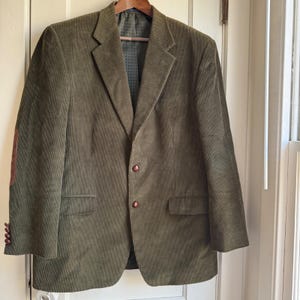 May include: Olive green corduroy blazer with brown elbow patches and matching buttons. The jacket is hanging on a wooden hanger against a white door. The blazer has a classic, tailored design.
