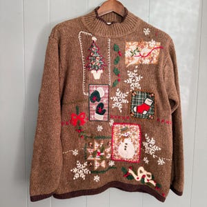 May include: A brown long-sleeve knit jumper featuring a mock neck and various holiday-themed appliqués. The design includes embroidered snowflakes, a Christmas tree, a snowman, a red stocking, and green mittens, all arranged in a patchwork style.