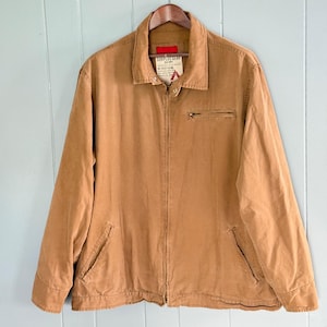 May include: A tan-colored jacket with a full zipper closure, a small chest pocket, and two lower pockets. The jacket has a collar and a tag that reads "SURPLUS GEAR". The jacket is hanging on a wooden hanger.