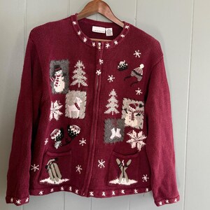 May include: A deep red zip-up knit cardigan featuring embroidered winter motifs including snowmen, pine trees, snowflakes, ice skates, a sled, and skis. The garment has a round neckline, long sleeves, and two front pockets with white accents.