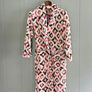 May include: A long, white dressing gown featuring a floral design of pink and red roses and green leaves. The gown has a shawl collar, two pockets, and white trim. It is hanging on a wooden hanger.