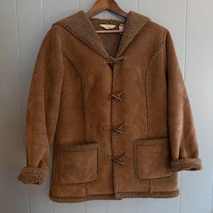 May include: A tan faux suede hooded coat with a brown sherpa fleece lining and trim. The coat features four wooden toggle closures down the front, two large patch pockets, and long sleeves with folded cuffs, all accented with visible stitching.