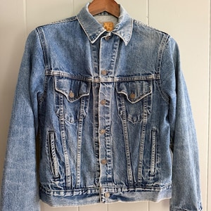 Puede incluir: Chaqueta vaquera azul claro con un diseño clásico. La chaqueta tiene cuello, cierre de botones en la parte delantera, dos bolsillos en el pecho y dos bolsillos laterales. La chaqueta está colgada en una percha de madera.