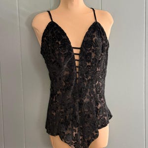 May include: Black bodysuit with a floral pattern. The lingerie features a deep V-neckline and thin spaghetti straps. The front has a series of vertical straps. The fabric has a velvet-like texture.
