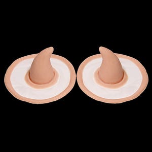 Puede incluir: Dos juegos de nariz y sombrero de bruja protésicos. Cada juego presenta una nariz curva de color marrón claro y una base de sombrero blanca y beige. Los artículos están sobre un fondo negro.