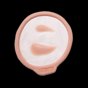 Puede incluir: Una pieza de maquillaje protésico con dos formas curvas color carne sobre una superficie blanca, dentro de un marco circular color piel. La marca "KEVEX" es visible. Diseñado para efectos especiales o uso teatral.