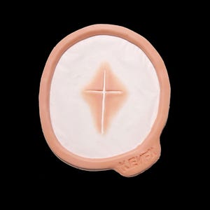 Puede incluir: Una herida protésica con un borde de tono de piel claro y un centro blanco. En el centro es visible una incisión en forma de cruz, con un tono marrón rojizo más oscuro. La palabra "KEVEN" está impresa en el lateral.