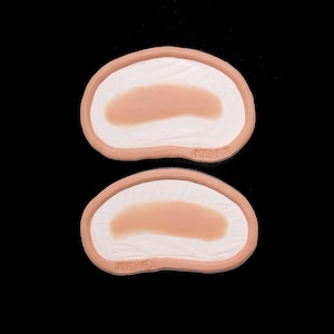 Puede incluir: Dos piezas de piel protésica, cada una con un centro marrón claro y un interior blanco, se muestran sobre un fondo negro. Los bordes son de color marrón más oscuro. La palabra "KENEX" está impresa en la parte inferior de cada pieza.