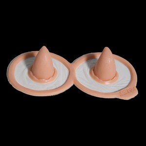 May include: A pair of connected, flesh-toned silicone nipple covers featuring a smooth, conical center and a wide, circular base. The surface has a matte finish with a subtle, radiating texture around the central point. The word LEIPA is embossed.