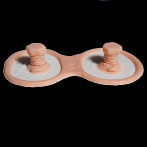 May include: A peach-colored, figure-eight shaped silicone device featuring two raised, threaded bolt-like protrusions on each side. The center bridge of the object is embossed with the text KEIR. The base of each circular section is white.