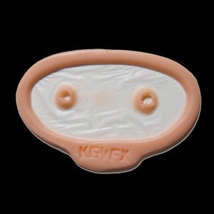May include: An oval-shaped prosthetic piece with a peach-toned border and a textured white center. The center features two small circular indentations. The word KEVEX is embossed in the peach-colored border at the bottom of the oval shape.