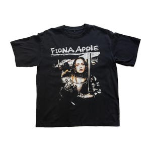May include: Black t-shirt featuring the text "Fiona Apple" in white, above a graphic of a woman in armor holding a sword. The image has a vintage, distressed look.