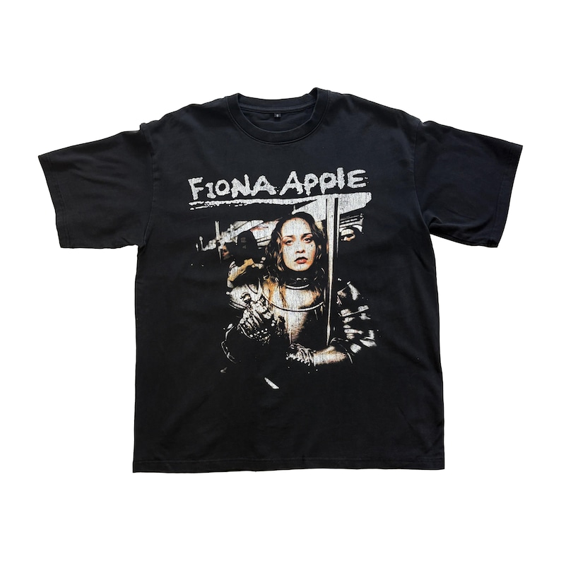 May include: Black t-shirt featuring the text "Fiona Apple" in white, above a graphic of a woman in armor holding a sword. The image has a vintage, distressed look.