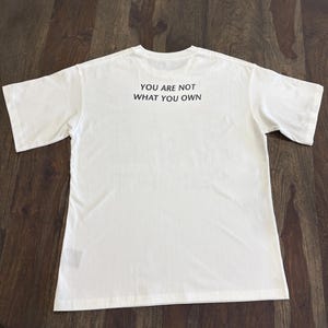 May include: A white t-shirt with the text "YOU ARE NOT WHAT YOU OWN" printed in black. The shirt is laid flat on a wooden surface, showcasing the back of the garment.