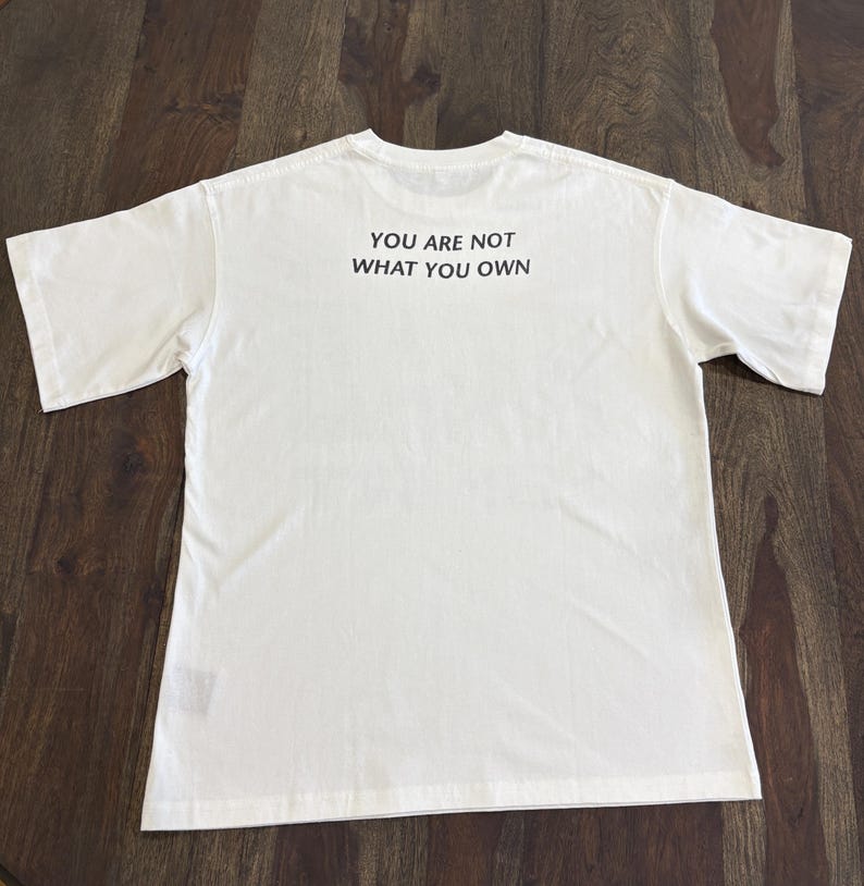 May include: A white t-shirt with the text "YOU ARE NOT WHAT YOU OWN" printed in black. The shirt is laid flat on a wooden surface, showcasing the back of the garment.
