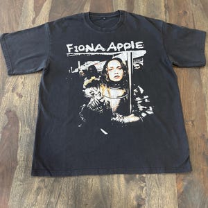 May include: Black t-shirt featuring the text "Fiona Apple" above a black and white graphic of a woman in armor. The graphic is a portrait of a woman with long hair and a stern expression.