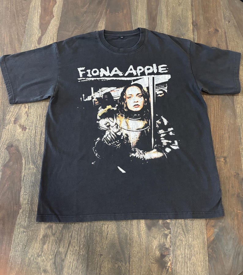 May include: Black t-shirt featuring the text "Fiona Apple" above a black and white graphic of a woman in armor. The graphic is a portrait of a woman with long hair and a stern expression.