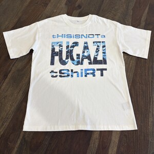 May include: A white t-shirt with the text "tHISISNOTa FUGazi tShirt" in blue and black. The t-shirt is made of a soft material and has short sleeves and a crew neck.