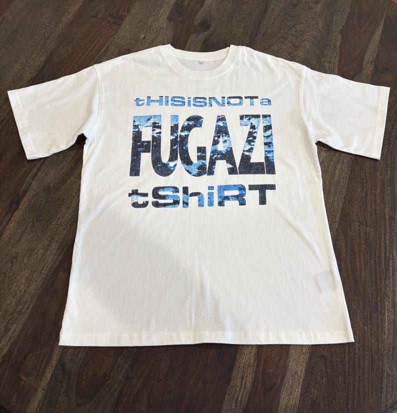 May include: A white t-shirt with the text "tHISISNOTa FUGazi tShirt" in blue and black. The t-shirt is made of a soft material and has short sleeves and a crew neck.