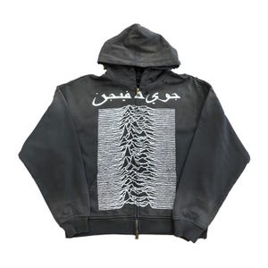 Puede incluir: Una sudadera con capucha con cremallera gris carbón con capucha. La parte delantera presenta un gráfico blanco de un diseño similar a una montaña y texto árabe encima. La sudadera tiene una cremallera completa y mangas largas.