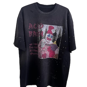 Peut inclure: T-shirt noir avec un motif vieilli d'un clown et les mots "ACID BATH" et "WHEN THE KIT STRING POPS". Le clown porte un chapeau rouge et une tenue rayée. Le t-shirt présente un aspect vintage et usé.