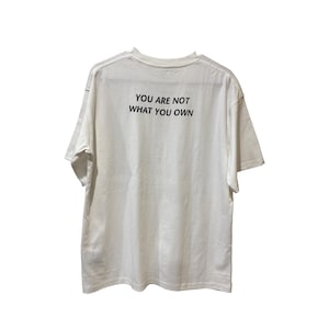May include: White t-shirt with the text "YOU ARE NOT WHAT YOU OWN" printed in black. The shirt has short sleeves and a crew neck. The fabric appears to be a soft cotton blend.