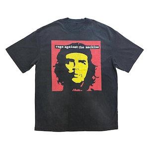 May include: Dark gray t-shirt featuring a yellow and black portrait on a red background. The text "rage against the machine" is printed above the portrait. The shirt is made of a soft material.