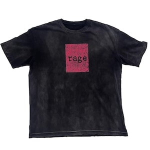 May include: Black t-shirt with a faded, vintage look. A distressed, rectangular pink graphic in the center features the word "rage" in a bold, black font. The shirt has a classic crew neck and short sleeves.