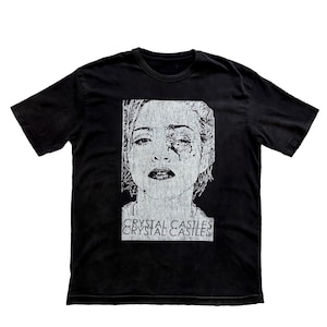 May include: Black t-shirt with a distressed white graphic of a woman's face and a tear. The text "CRYSTAL CASTLES" is printed below the image in a matching distressed font. The shirt has short sleeves and a crew neck.